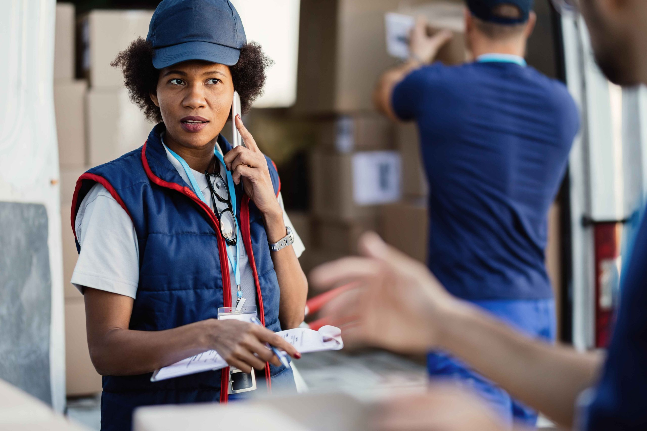 black-female-deliverer-communicating-with-coworker-while-talking-mobile-phone-organizing-package-delivery